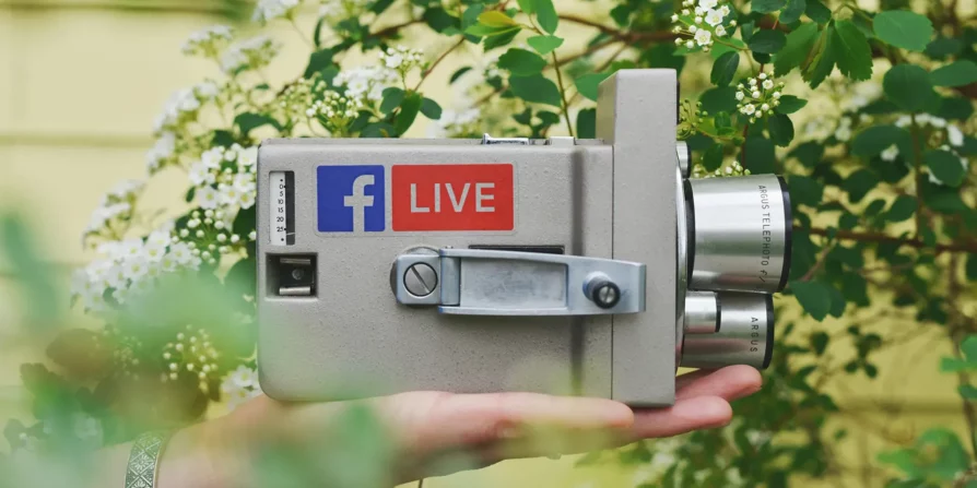 Una mano tiene una videocamera vintage con adesivi di Facebook e LIVE, mentre è circondata da fiori verdi e bianchi