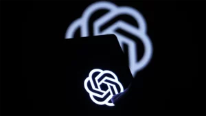 Logo di ChatGPT visibile su uno schermo in un ambiente scuro, con una luce blu che evidenzia il simbolo, mentre sullo sfondo si intravede una versione sfocata dello stesso logo