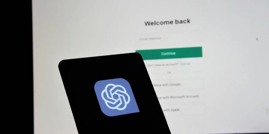Una persona tiene un telefono cellulare con il logo di ChatGPT in primo piano, mentre sullo sfondo si vede una schermata di accesso che chiede di inserire l'indirizzo email per continuare