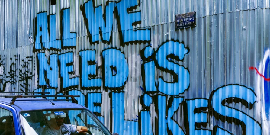 Graffiti su una parete di lamiera che dice "ALL WE NEED IS LIKES" con un'auto blu che passa davanti