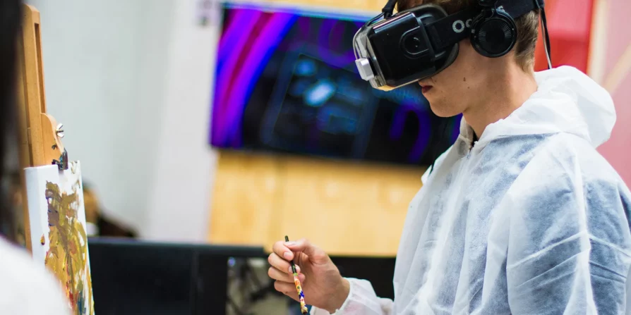 Persona che indossa un visore VR e cuffie mentre dipinge su una tela, in uno studio artistico, con una tuta protettiva bianca