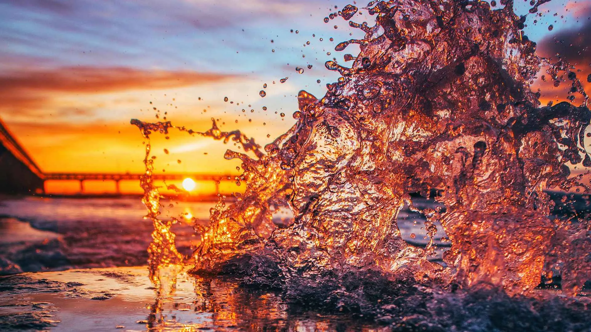 Un'immagine al tramonto con l'acqua che schizza dalla riva del mare. La luce del sole si riflette sull'acqua, creando una scena di colori caldi con il cielo che sfuma tra il giallo, arancione e il blu. In lontananza si vede un ponte.