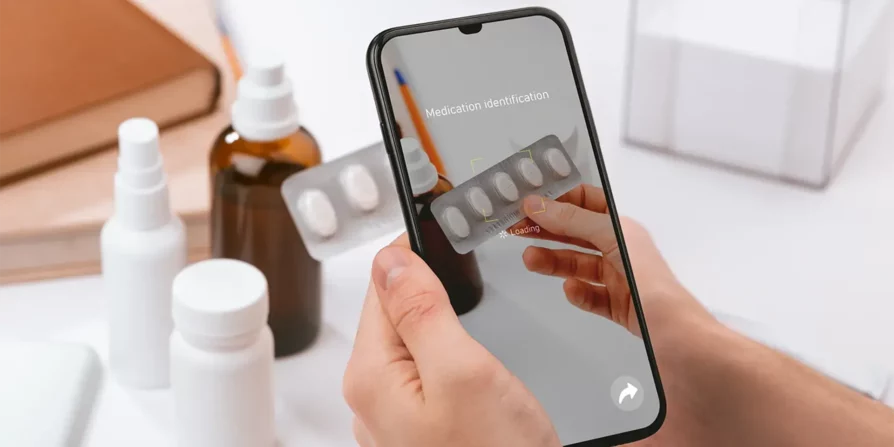 Una persona sta utilizzando uno smartphone per identificare un medicinale attraverso la fotocamera. L'applicazione sullo schermo del telefono mostra un caricamento mentre analizza un blister di pillole. Sullo sfondo, ci sono diversi contenitori di medicina e un quaderno